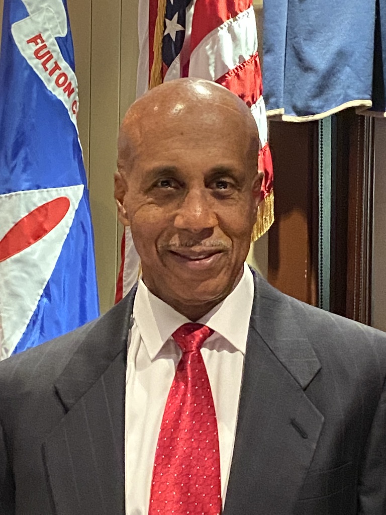 Portrait of Captain Paul Thompson in a dark suit and red tie in front of American and county flags